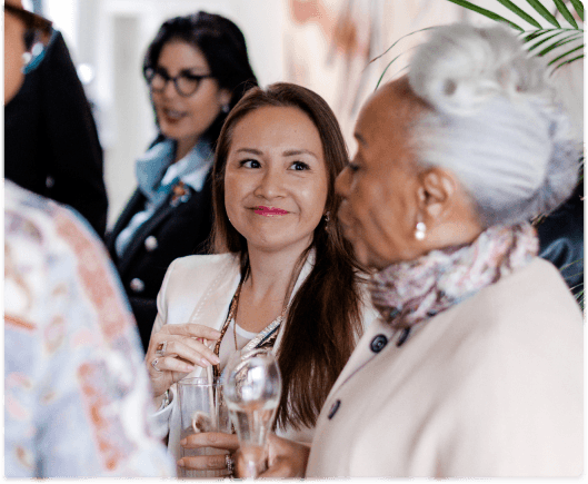 A group of women holding drinks and engaging in conversation with the camera focused on one woman listening and smiling
