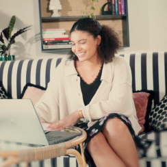 a smiling woman seated on a sofa working on a laptop